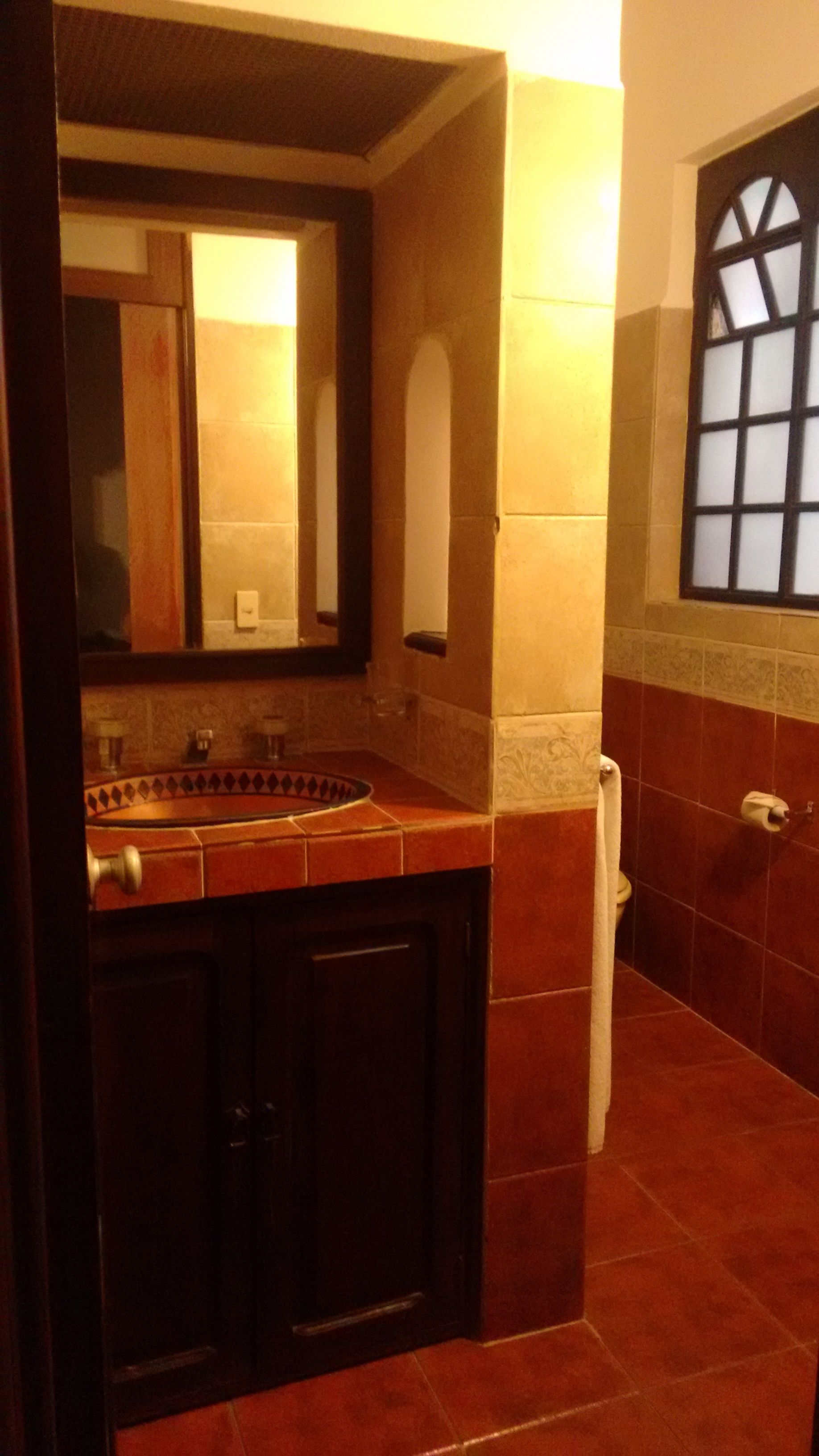 Un baño con un mueble de madera oscura, un lavabo y un espejo. Hay un perchero en la pared y una ventana con diseño de cuadrícula.