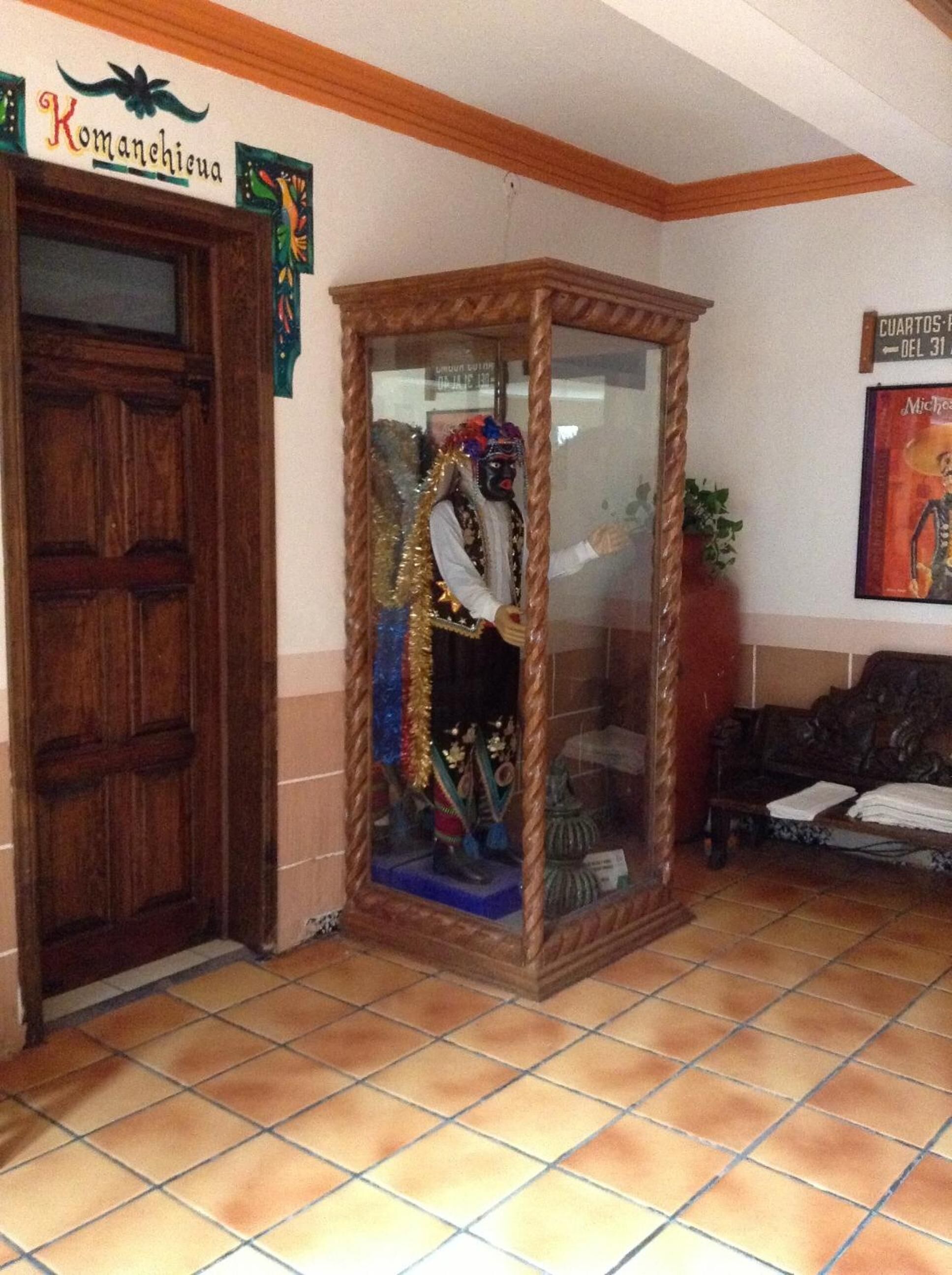 A room with a wooden door, a glass display case with a statue, a sign reading "Komanchicua", and a poster on the wall.