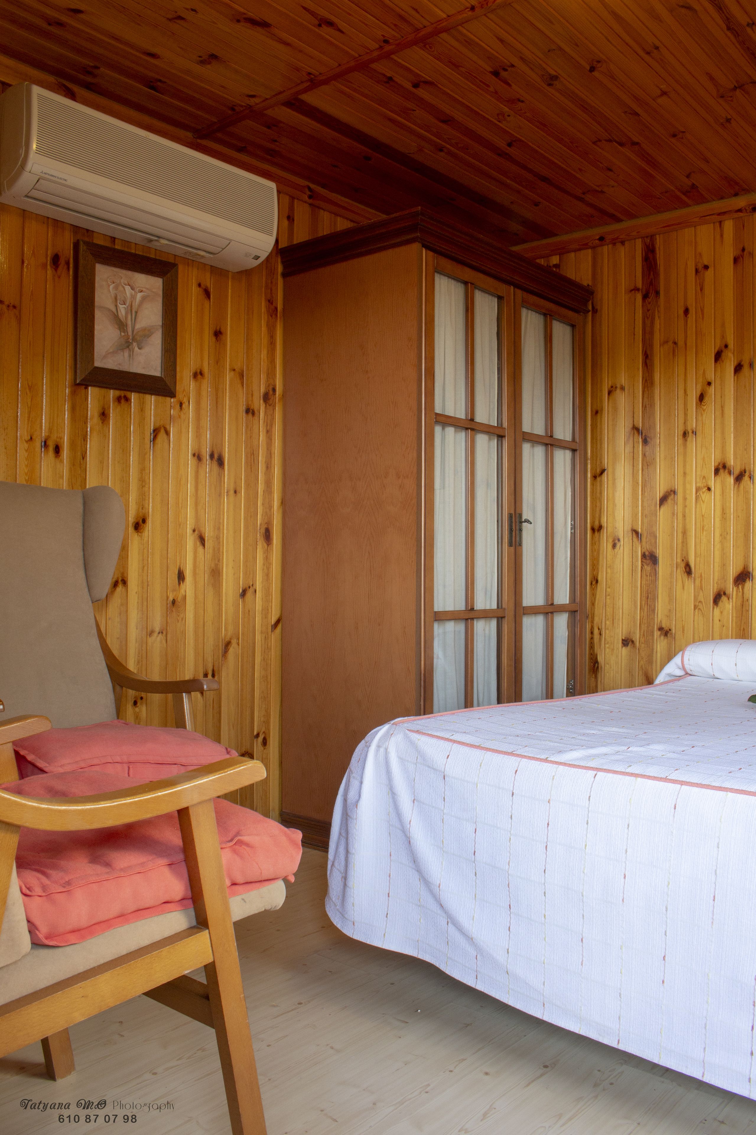 Une pièce aux murs lambrés, avec un lit, une chaise dotée d’un coussin rose et un climatiseur fixé au mur.