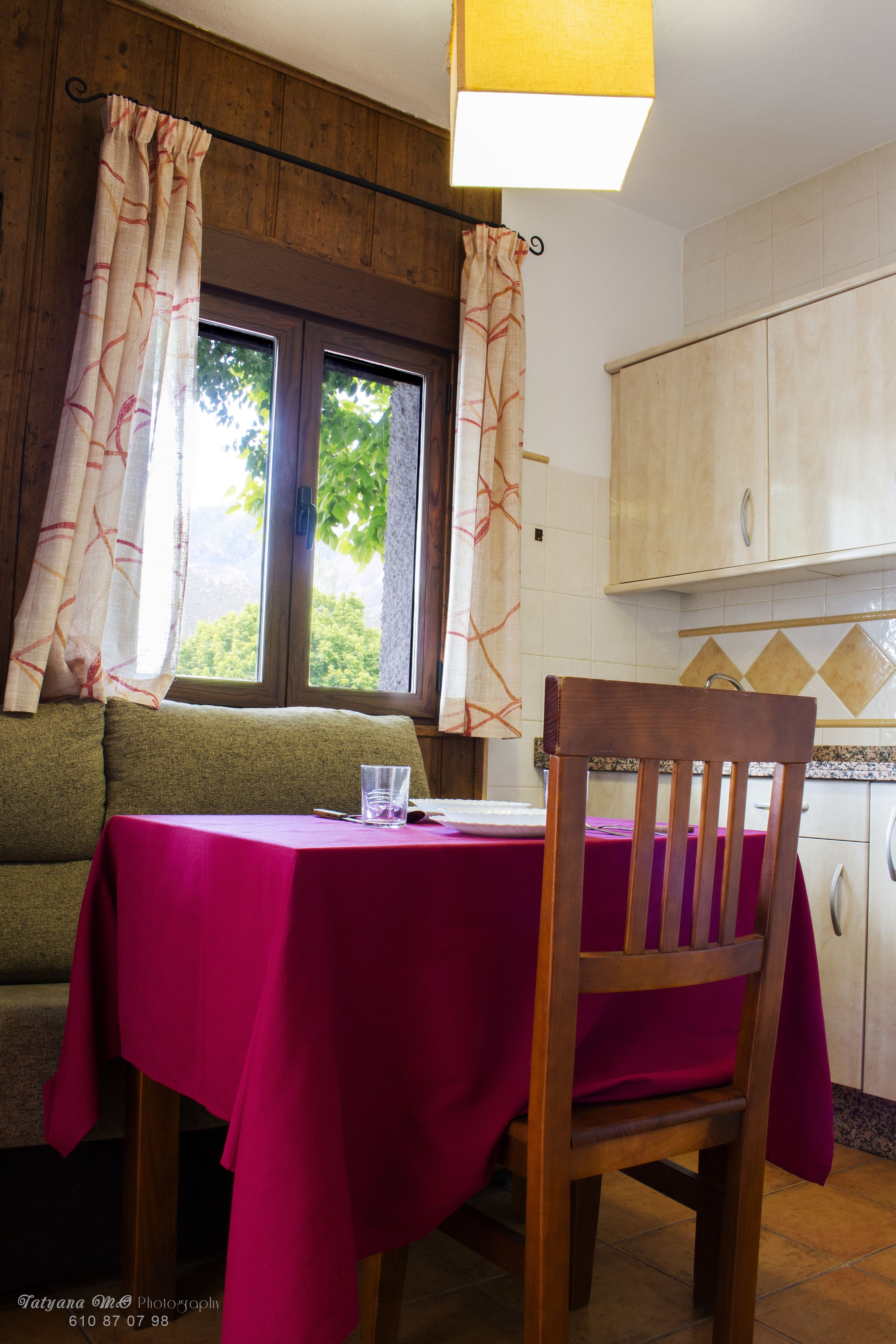 Un área de comedor con una mesa dispuesta para dos, una silla de madera, un sofá y una ventana con cortinas.