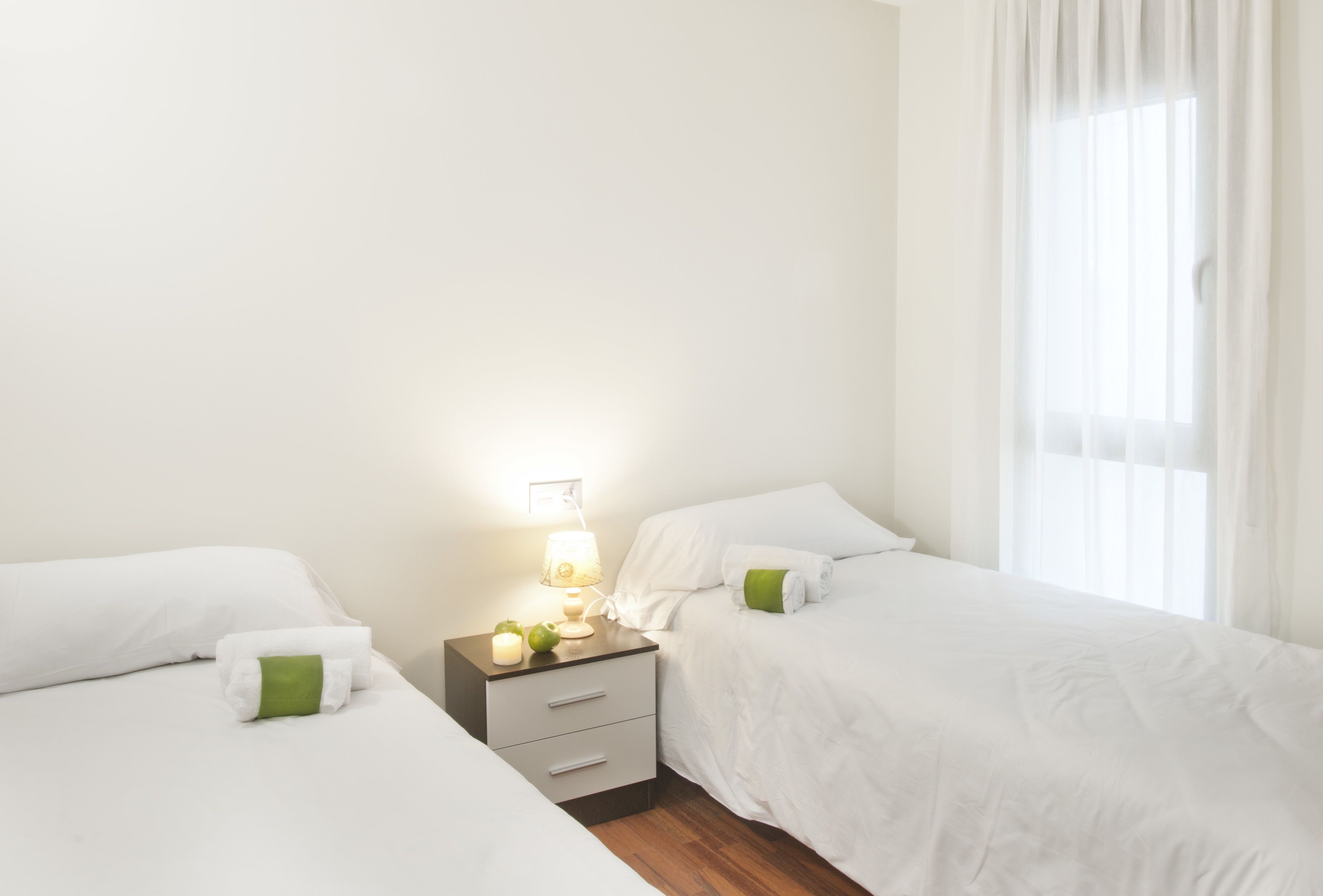 A hotel room with two beds, a nightstand with a lamp, and a window with sheer curtains.