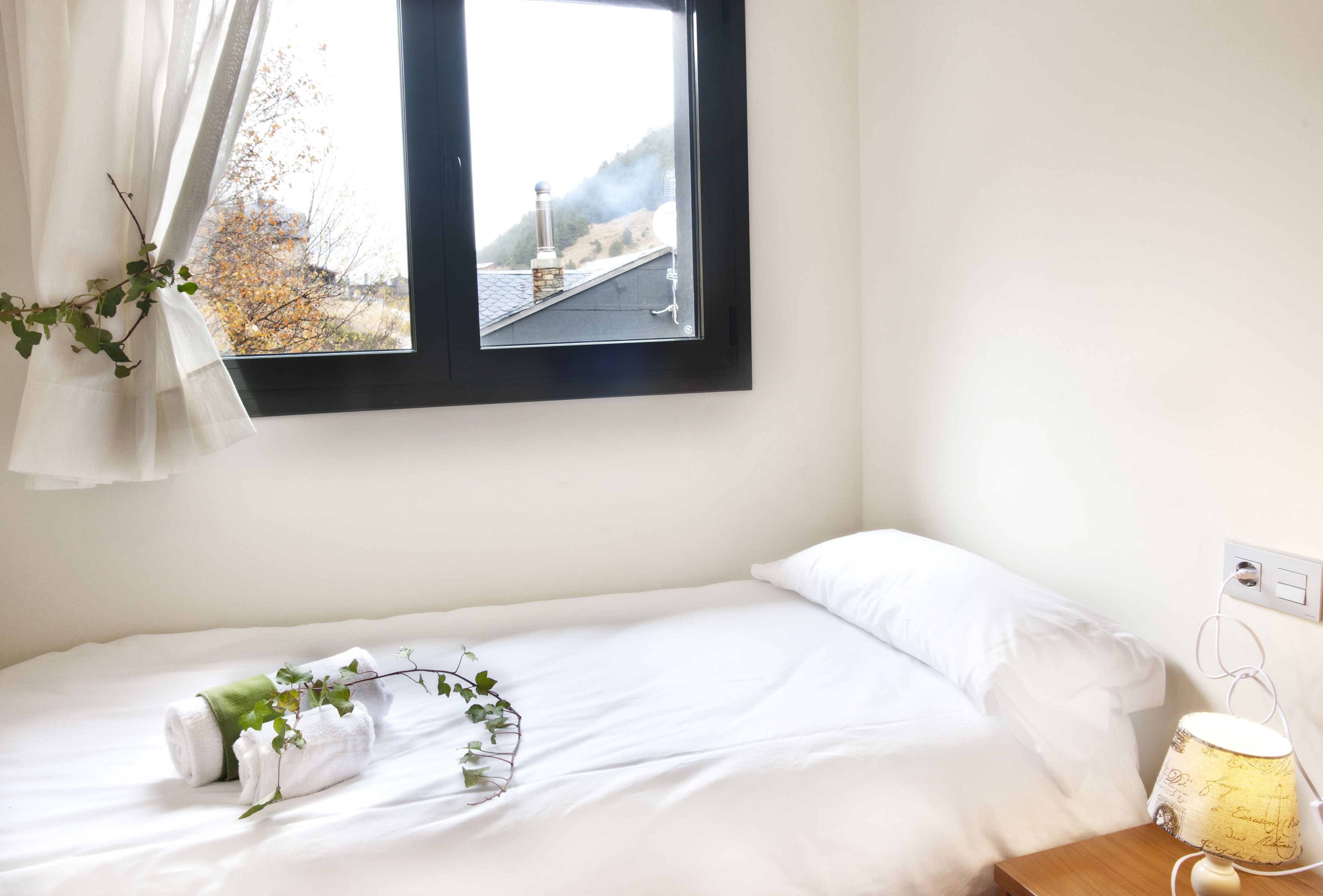 A neatly made bed with white linens, a bedside lamp, and a window with a view of a building and trees.