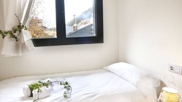 A neatly made bed with white linens, a bedside lamp, and a window with a view of a building and trees.