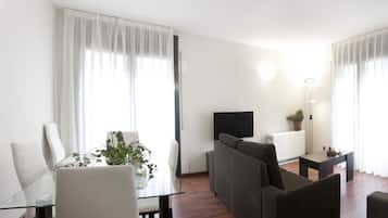 Un salon moderne avec une table à manger en verre, des chaises blanches, un canapé foncé, un téléviseur et une petite table d’appoint agrémentée d’une plante.
