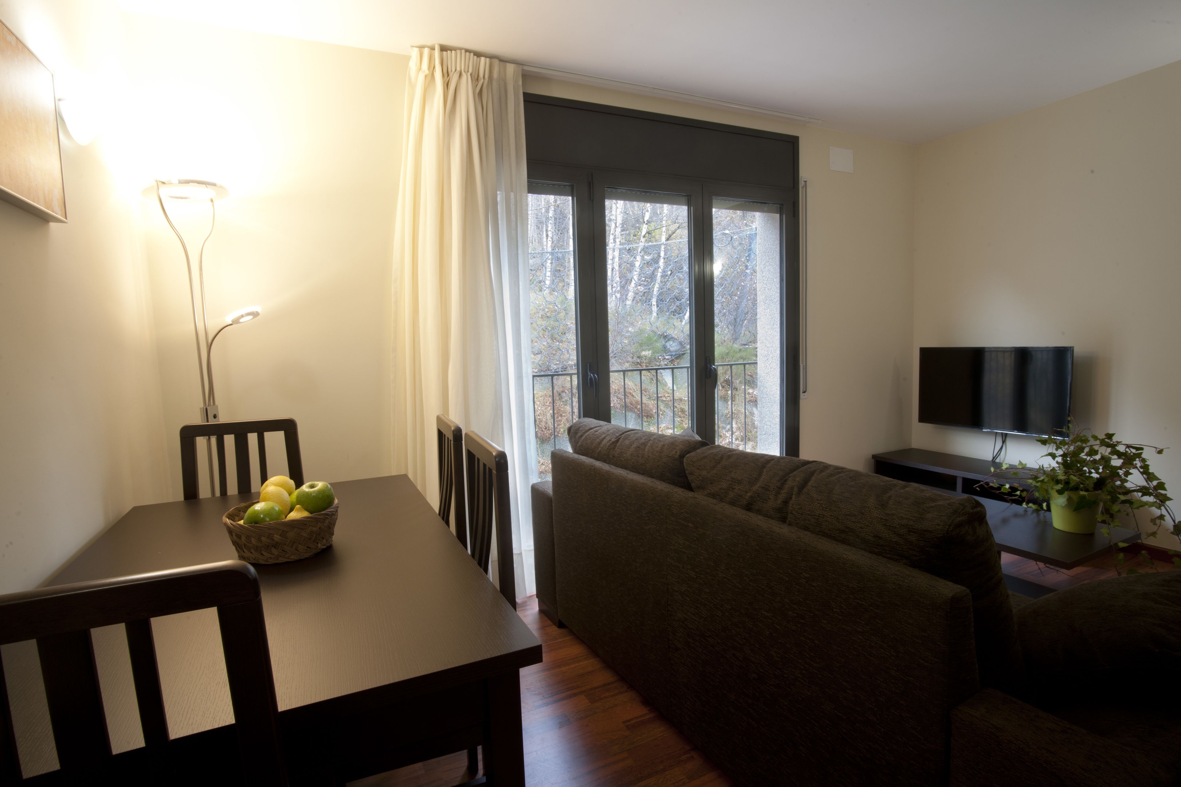 A living room with a dining table, chairs, a sofa, a TV, and a window with a view of trees.