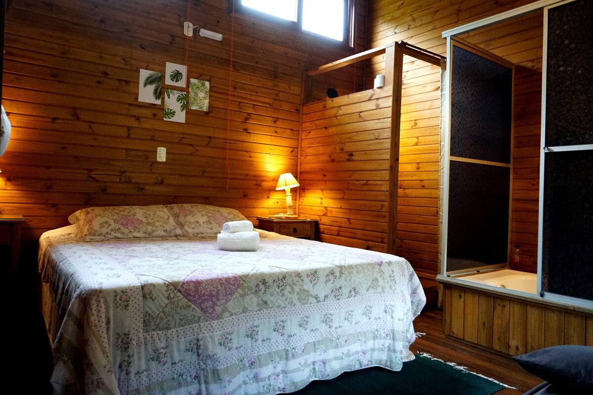 Quarto de cabana de madeira com uma cama, uma mesinha de cabeceira, uma luminária e uma janela grande.
