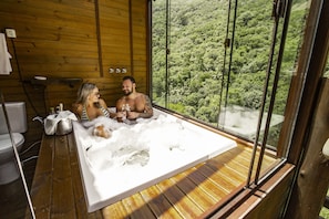 A wooden cabin with a large window overlooking a forest. Inside, there is a wooden deck and a hot tub with two people relaxing. - Cachoeira dos Borges Cabanas e Parque (Mampituba)