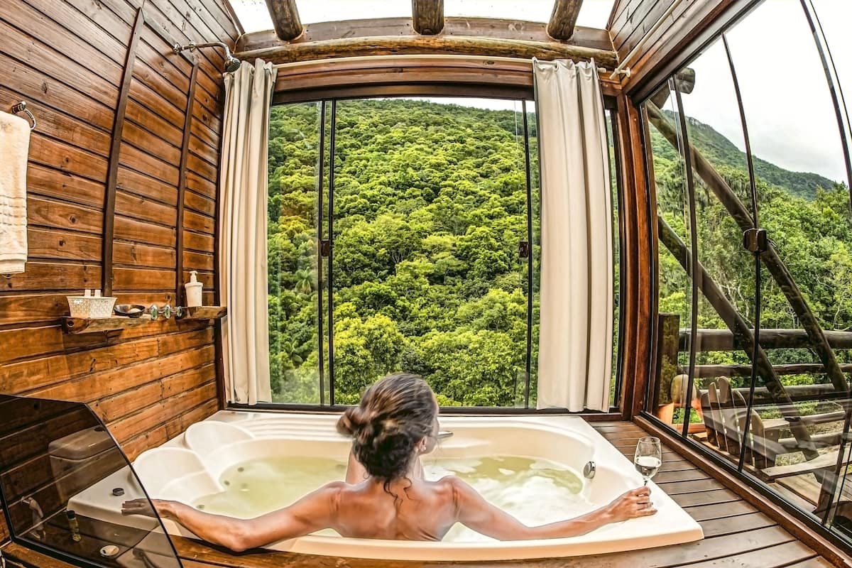 Uma pessoa relaxando em uma banheira de madeira com vista para uma encosta arborizada.