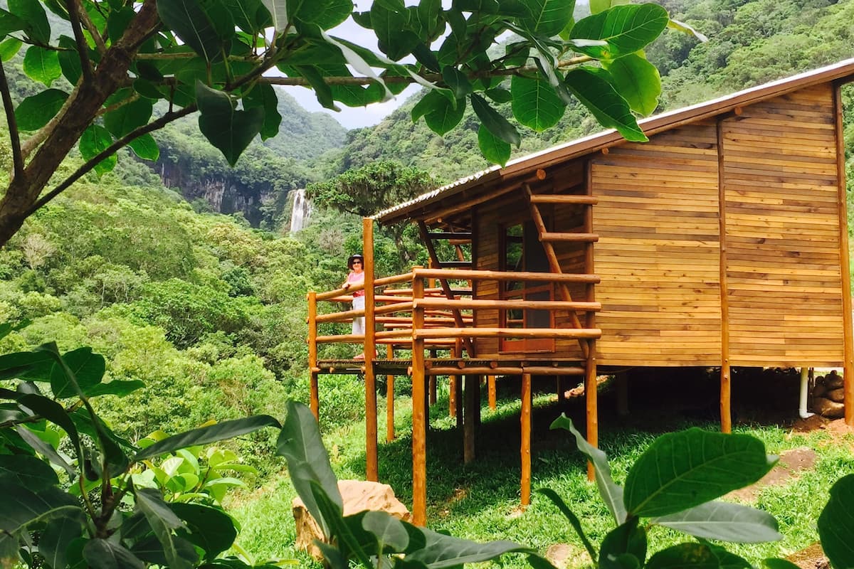 Uma cabana de madeira com um deck de madeira, rodeada por vegetação exuberante e um cenário montanhoso.