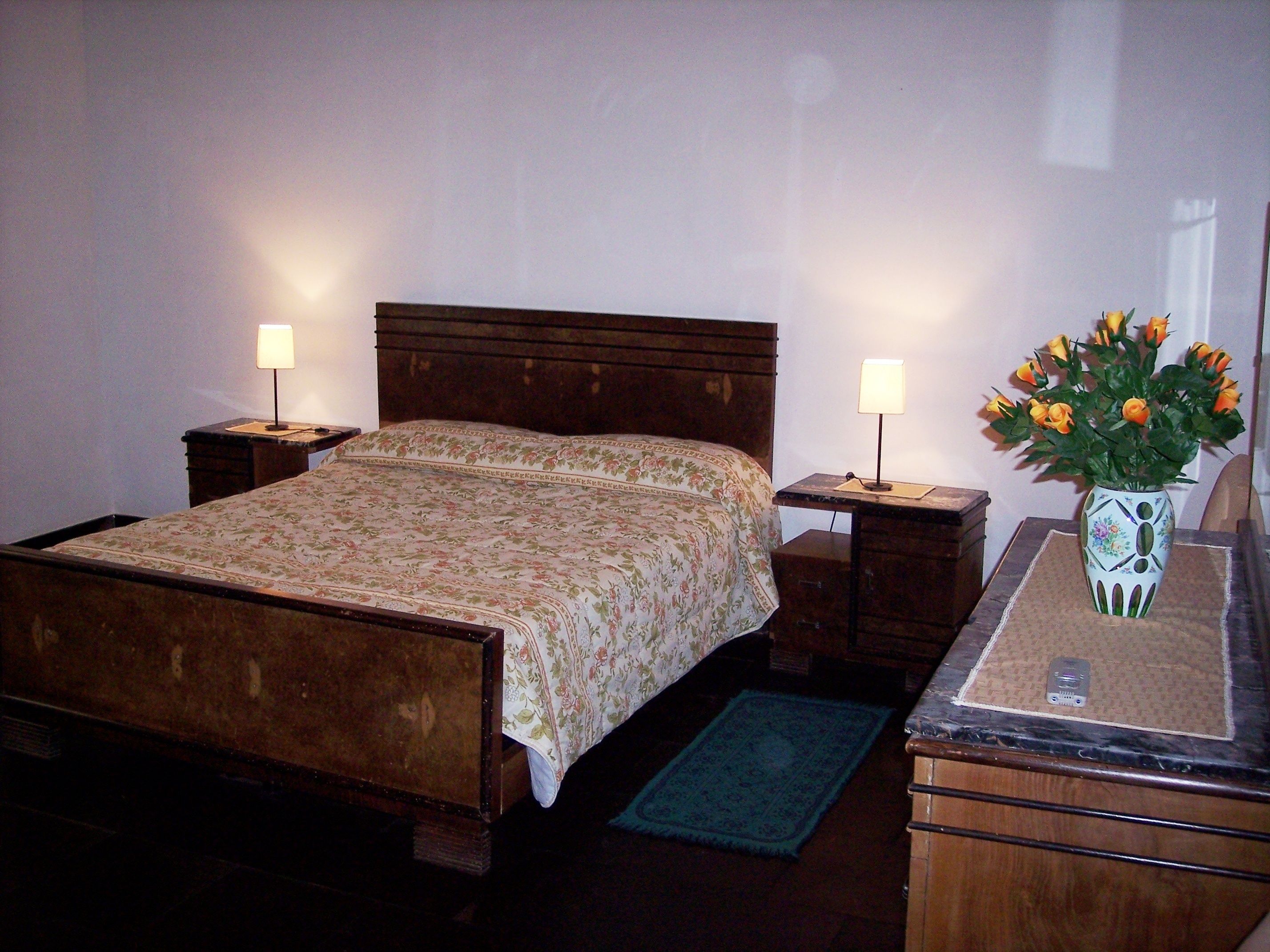 A bedroom with a wooden bed, two bedside tables with lamps, a nightstand, a dresser, a vase with flowers, and a blue rug.