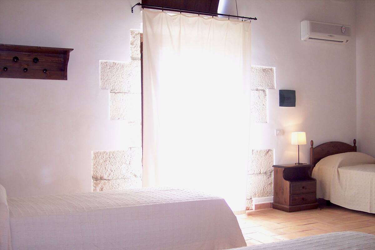 Quarto com duas camas, mesa de cabeceira com abajur, cabeceira de madeira e janela com cortina.
