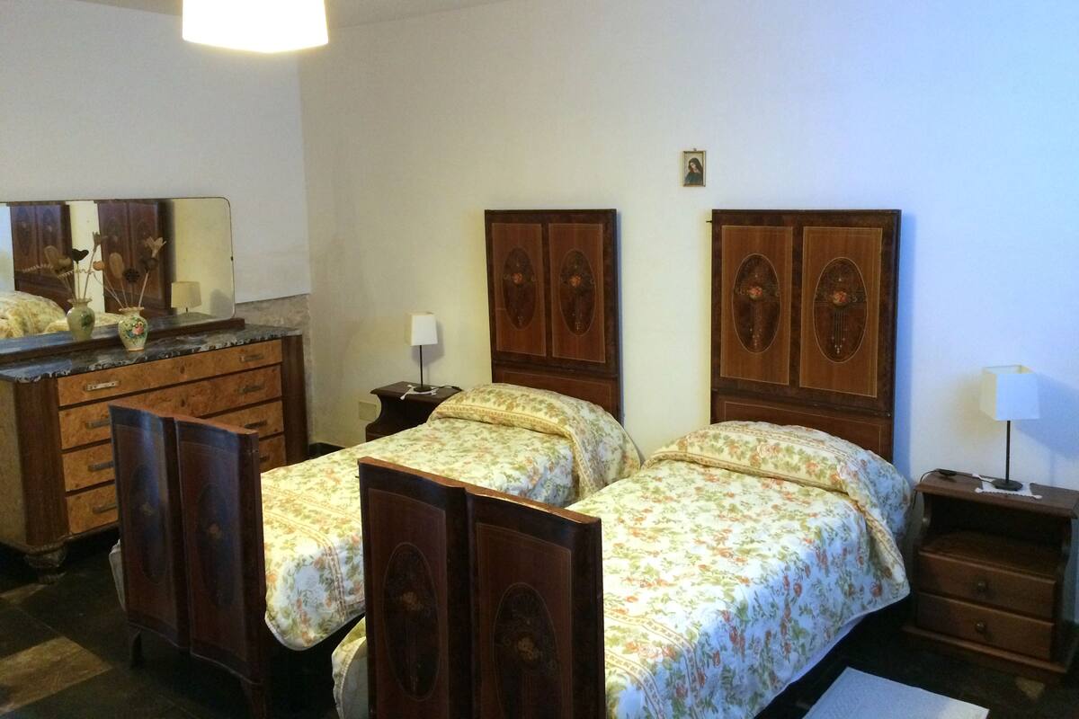 Quarto com duas camas de madeira, cômoda, espelho e mesa de cabeceira com abajur.