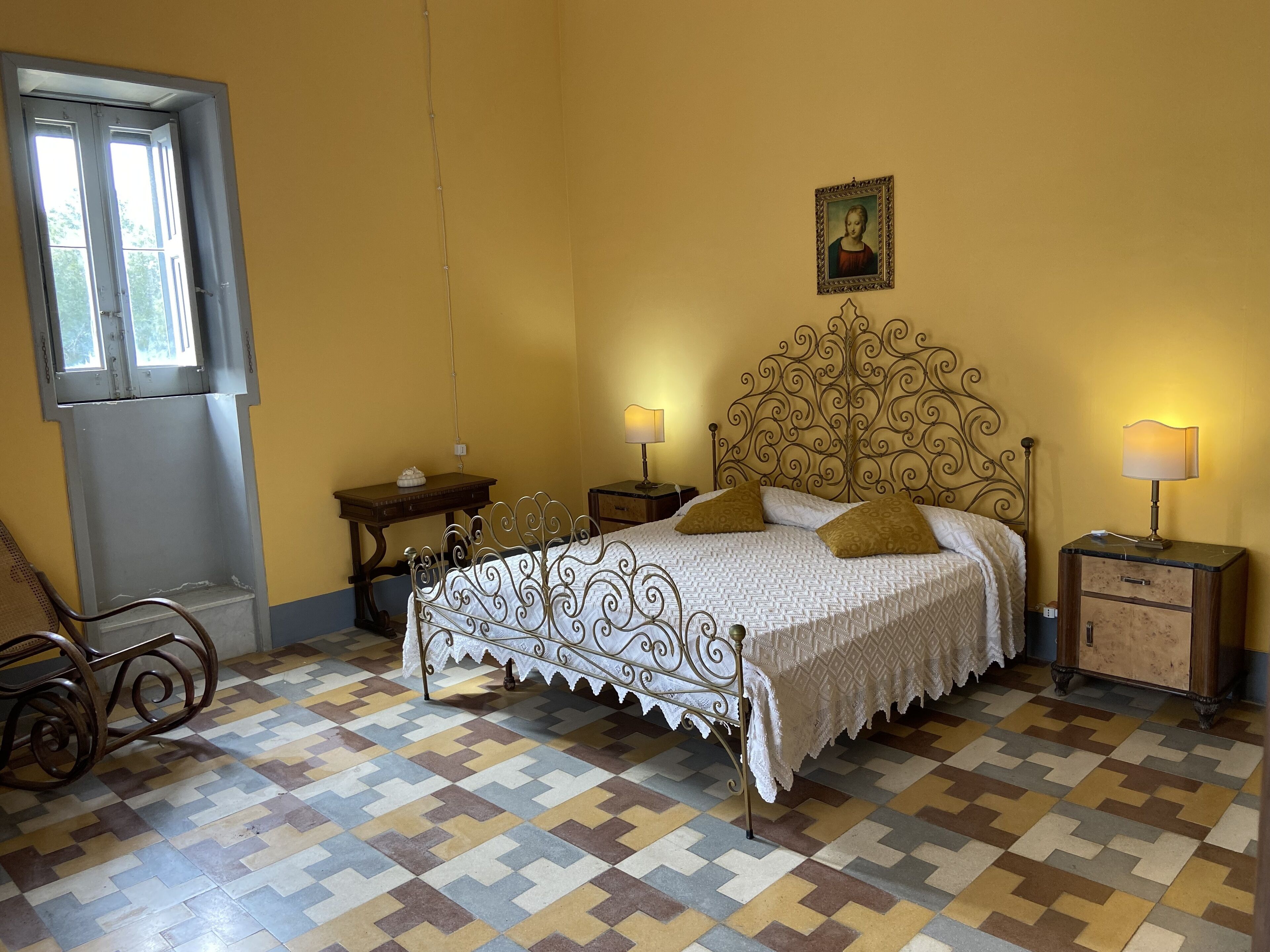 Une chambre avec un sol à motifs, un lit doté d’une tête de lit décorative, deux tables de chevet avec des lampes, un fauteuil et une fenêtre donnant sur un paysage agréable.