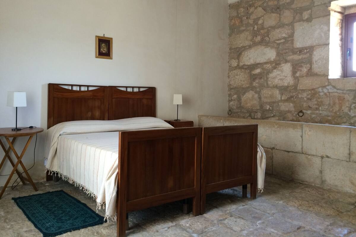 Quarto com cama de madeira, duas mesinhas de cabeceira, um tapete pequeno, parede de pedra e uma janela.