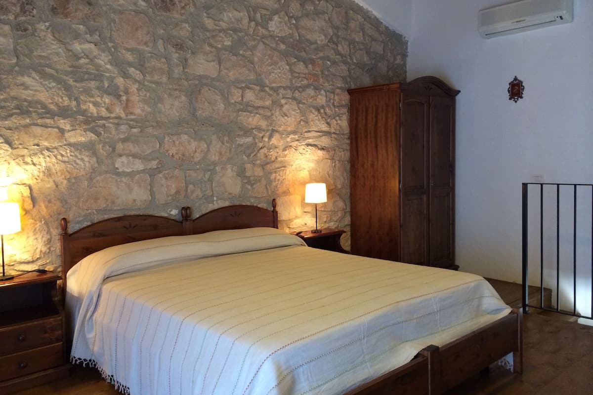 Quarto com parede de pedra, cama de madeira, mesas de cabeceira e guarda-roupa.