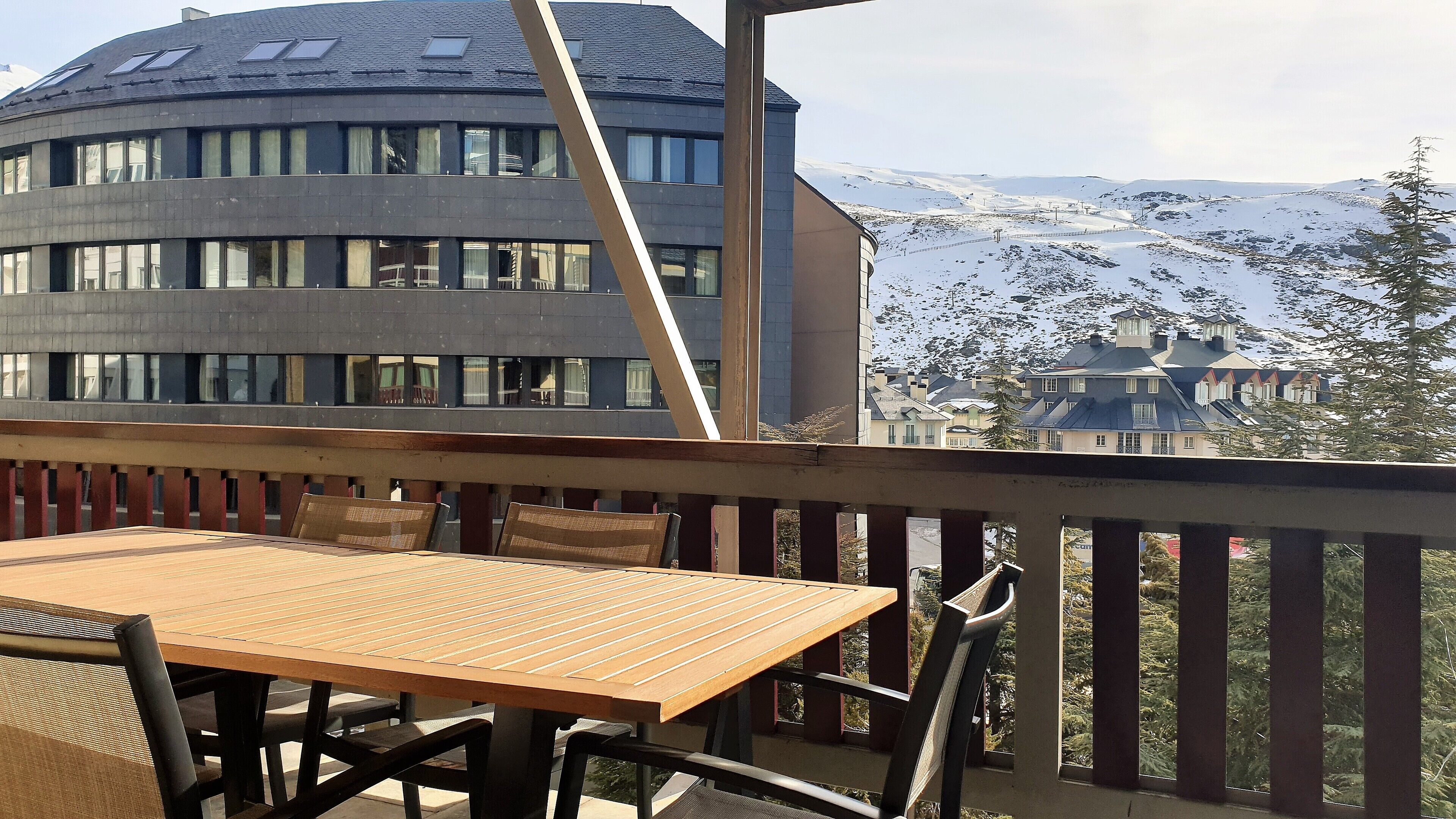 Une table et des chaises en bois sur un balcon extérieur, avec vue sur un immeuble moderne et des montagnes enneigées.