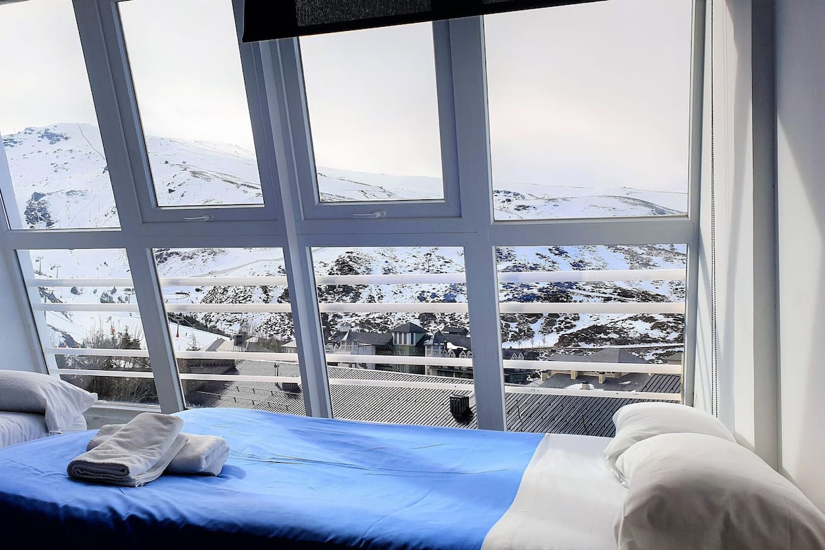 Quarto com grande janela oferecendo vista de uma paisagem montanhosa coberta de neve.