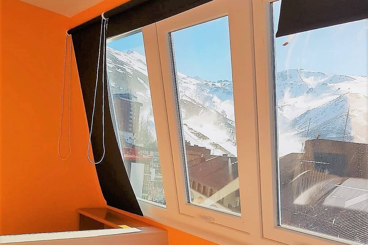Um quarto com cama, almofadas, janela com vista para montanhas cobertas de neve e cortina.