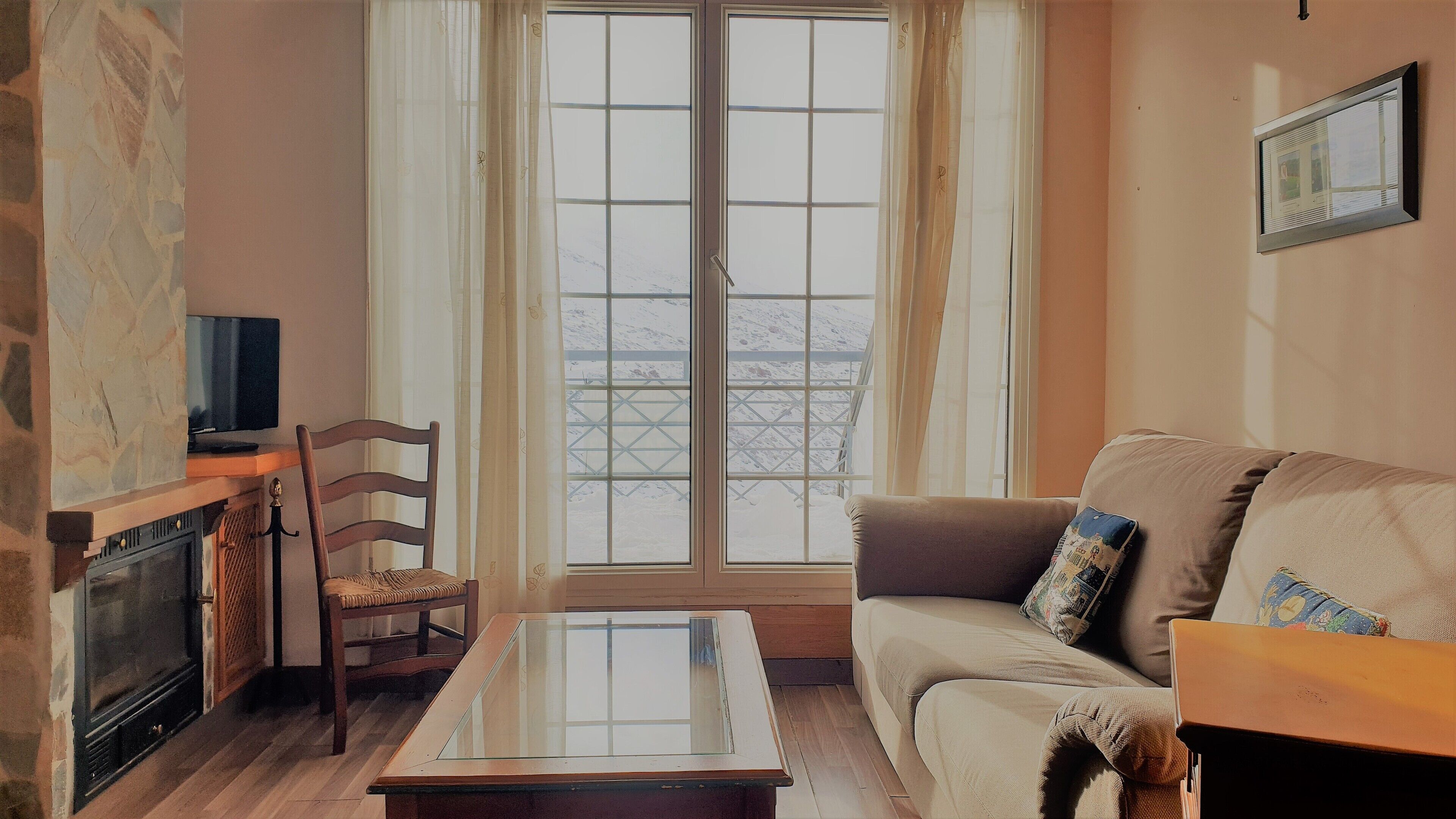 Una sala de estar con chimenea de piedra, una silla de madera, una mesa de centro con superficie de vidrio, un sofá color beige con cojines estampados y un televisor sobre un mueble de madera.