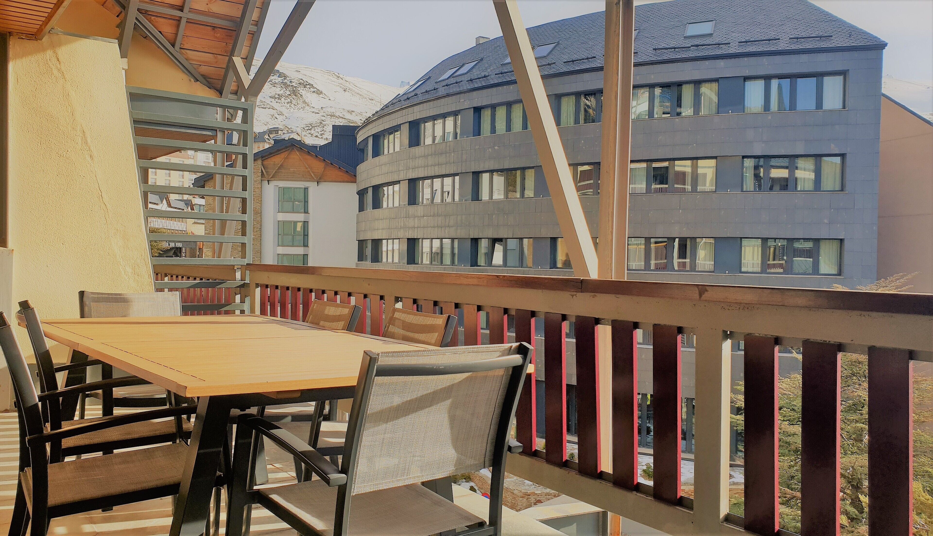 Un balcón con una mesa y sillas de madera, con vistas a un edificio moderno con múltiples ventanas y una montaña nevada al fondo.