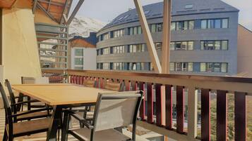 Un balcón con una mesa y sillas de madera, con vistas a un edificio moderno con múltiples ventanas y una montaña nevada al fondo.