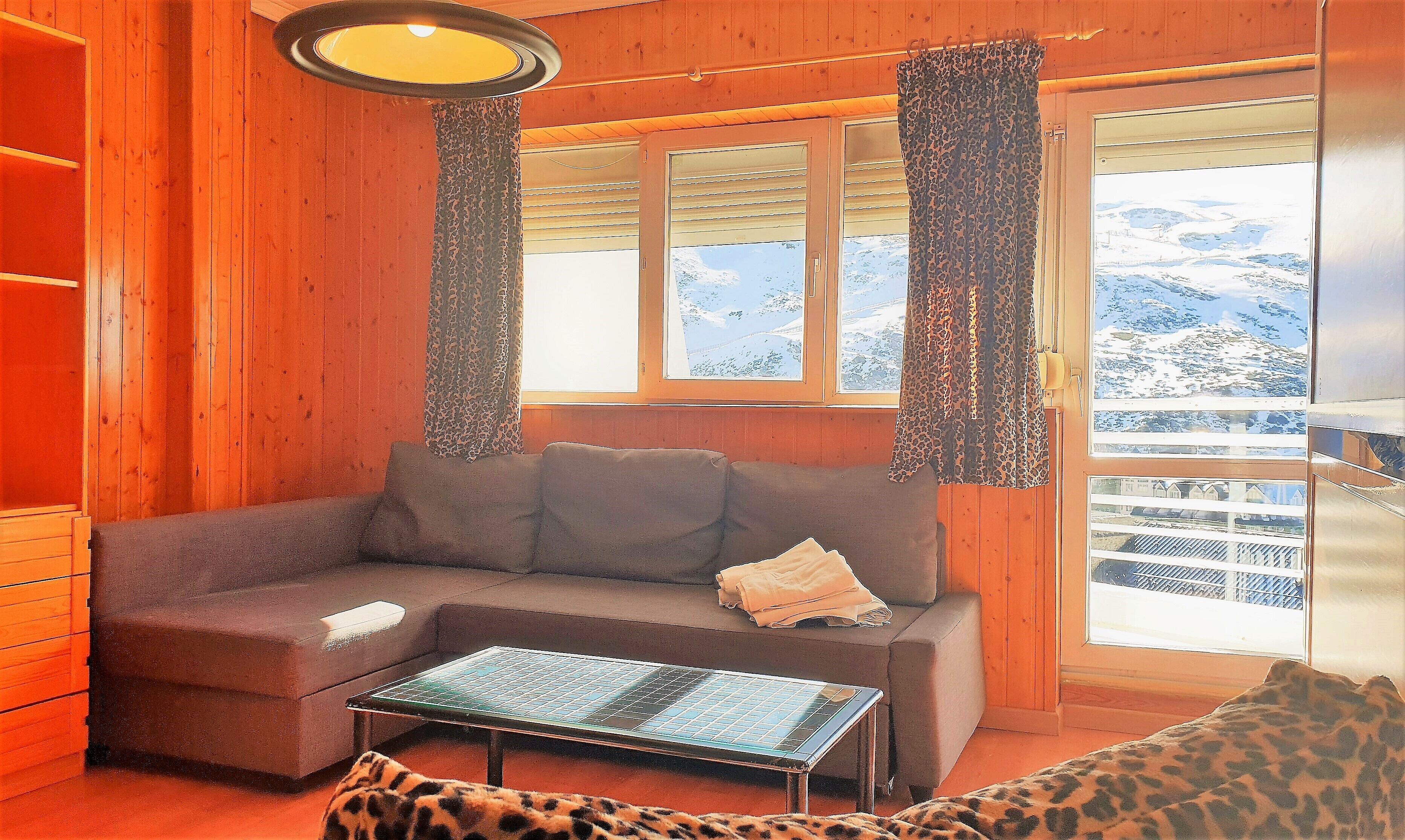 Interior acogedor de una cabaña con un sofá, una mesita y vistas a montañas nevadas a través de amplios ventanales.