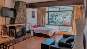 Habitación de hotel con chimenea de piedra, cama con almohadas rojas, televisor montado en la pared y un ventanal con vistas a un pueblo.