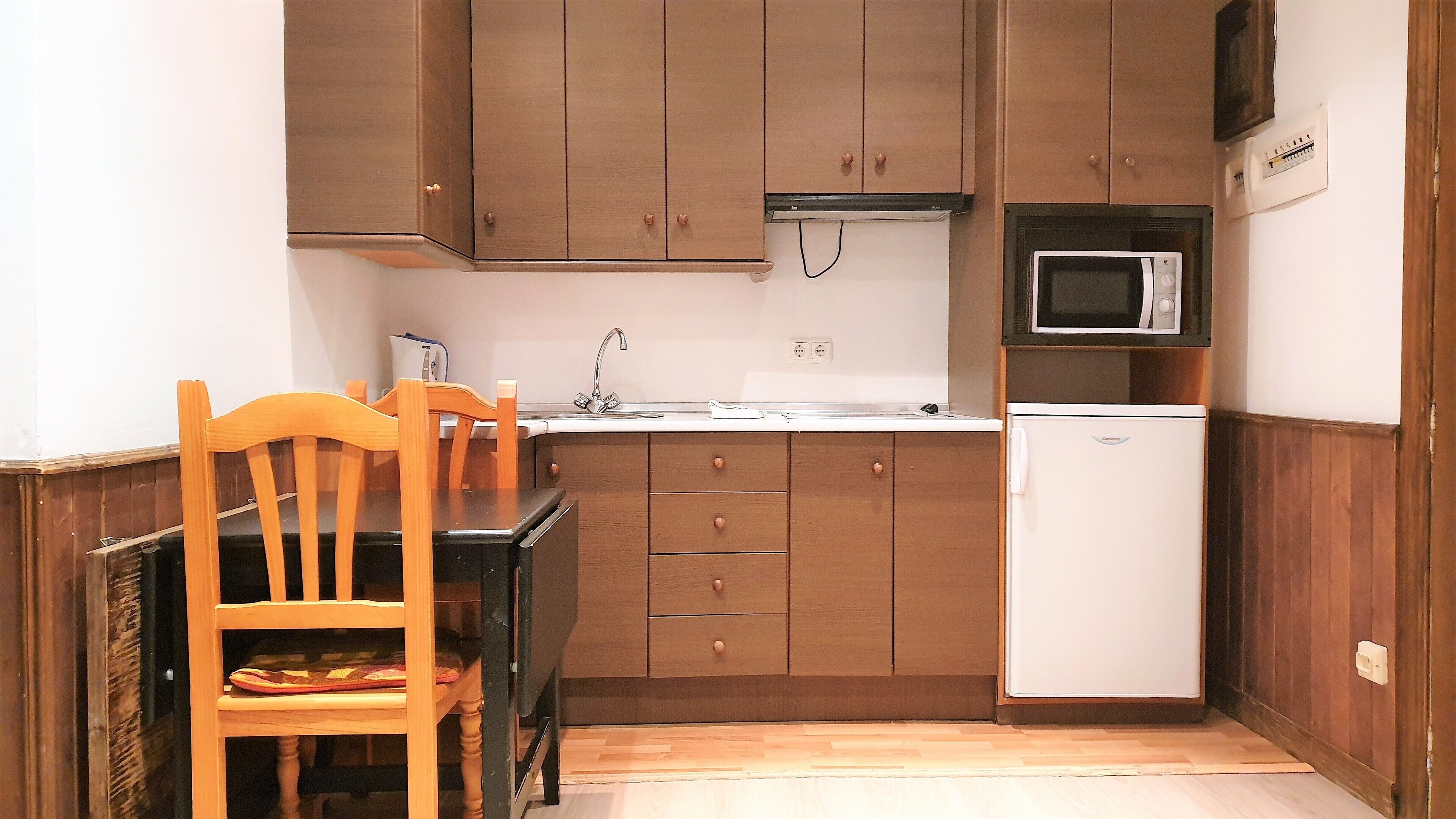 A compact kitchen with wooden cabinets, a white refrigerator, a microwave, and a dining table with chairs.