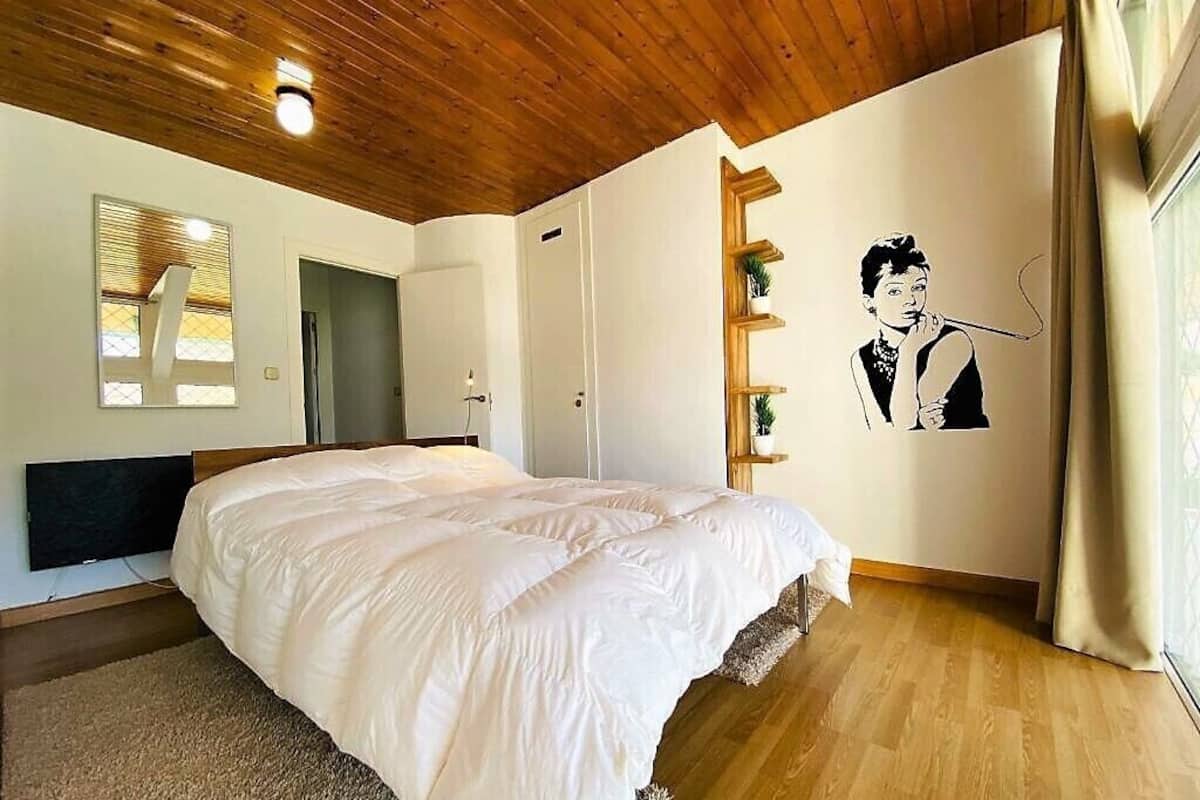 Um quarto com uma cama grande, teto de madeira, uma prateleira na parede com uma planta e um quadro de uma pessoa segurando um cachimbo.