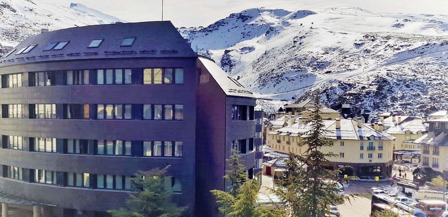 Un edificio moderno de varios pisos y grandes ventanales, rodeado de montañas nevadas.