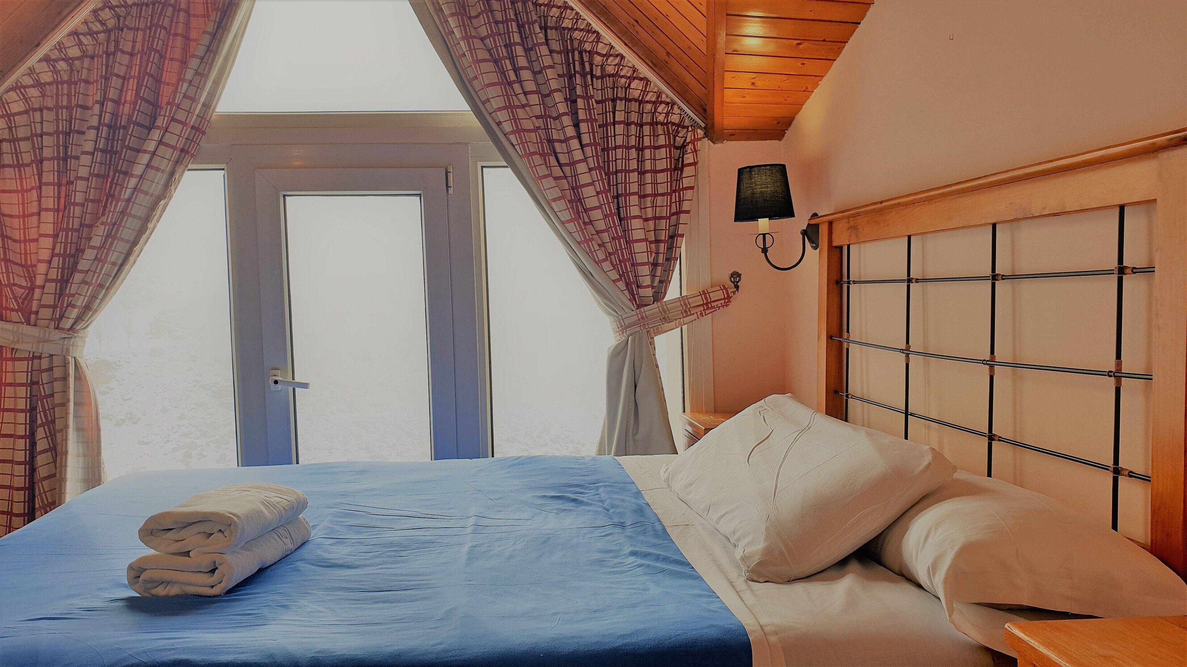 A bedroom with a bed, a nightstand, a wall-mounted lamp, and a window with curtains.