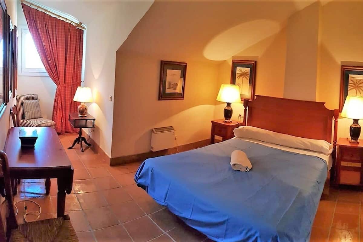 Quarto de hotel com cama, duas mesas de cabeceira com luminárias, uma cadeira, escrivaninha e janela com cortinas.