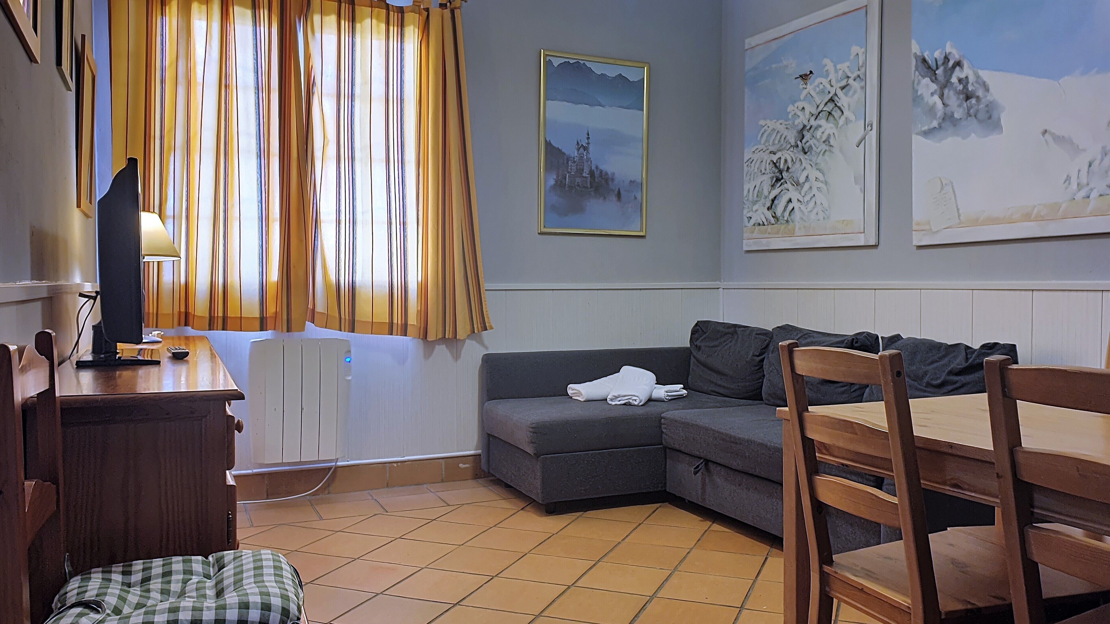 Een gezellige woonkamer met een hoekbank, een houten stoel, een tafeltje met een lamp en een radiator.