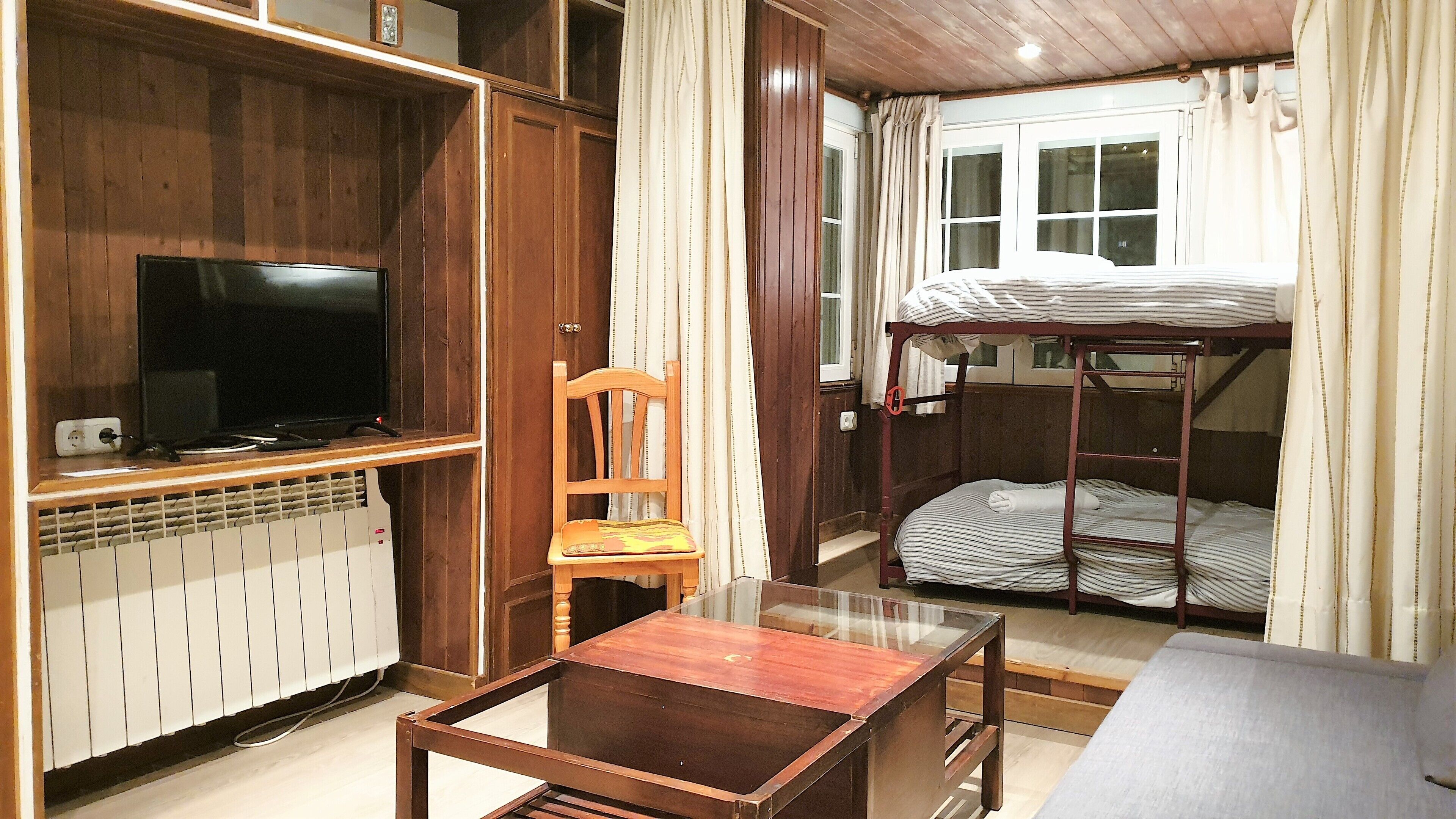 Una cama litera con ventana, un televisor en un estante de madera, una silla y una mesa en una habitación con paredes de madera y suelo de baldosas.