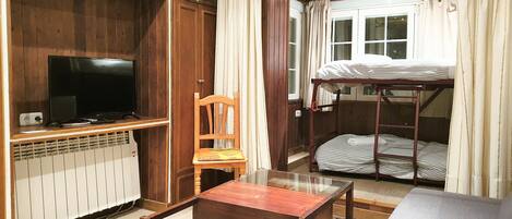 Una cama litera con ventana, un televisor en un estante de madera, una silla y una mesa en una habitación con paredes de madera y suelo de baldosas.