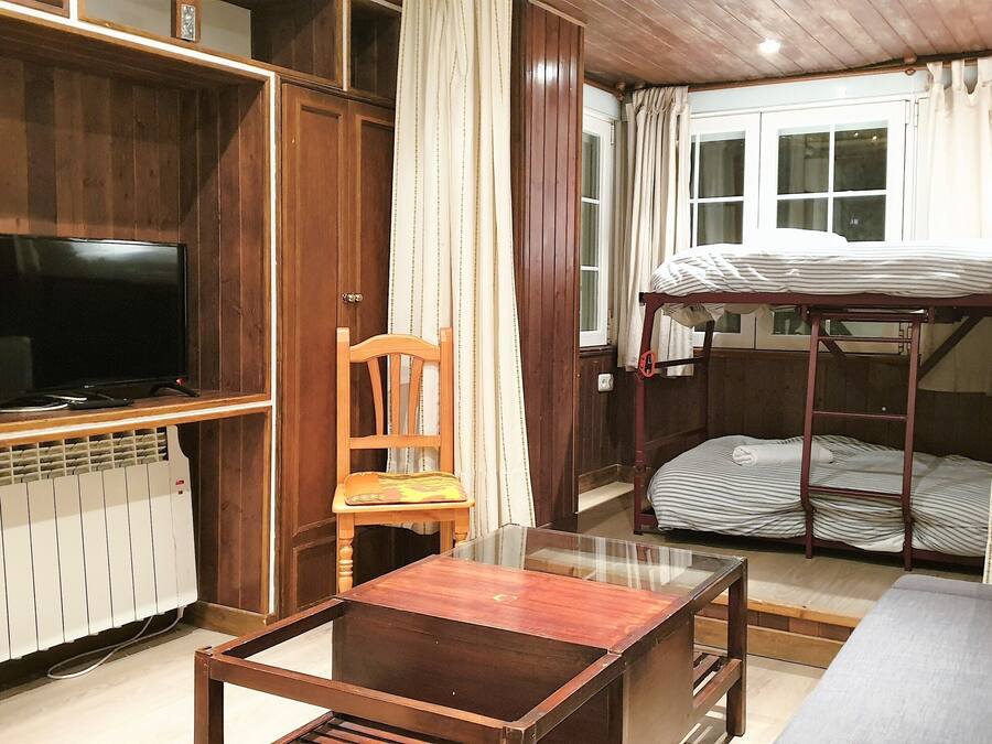 Una cama litera con ventana, un televisor en un estante de madera, una silla y una mesa en una habitación con paredes de madera y suelo de baldosas.