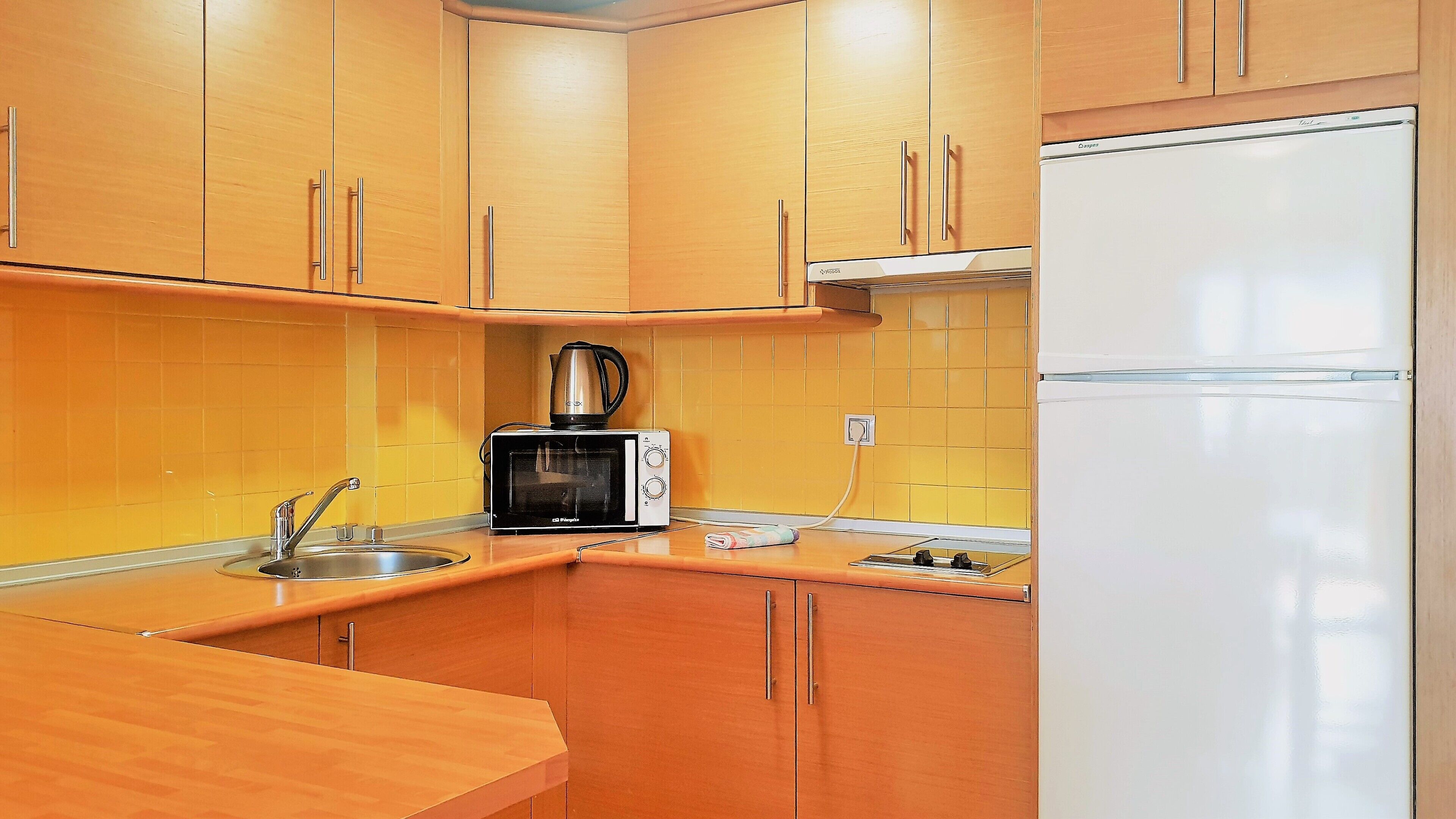 A kitchen with wooden cabinets, a microwave, a kettle, and a refrigerator.