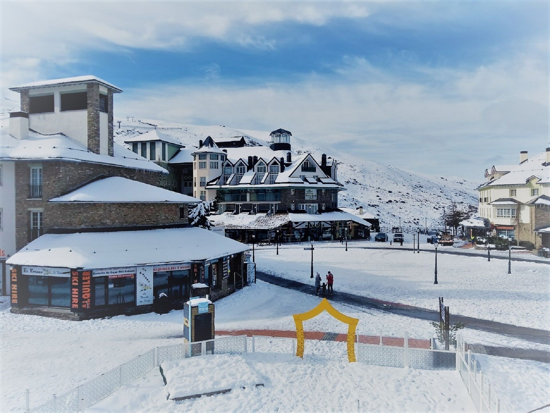 A snow-covered building with a ski school sign, a ski hire shop, and a mountain backdrop.