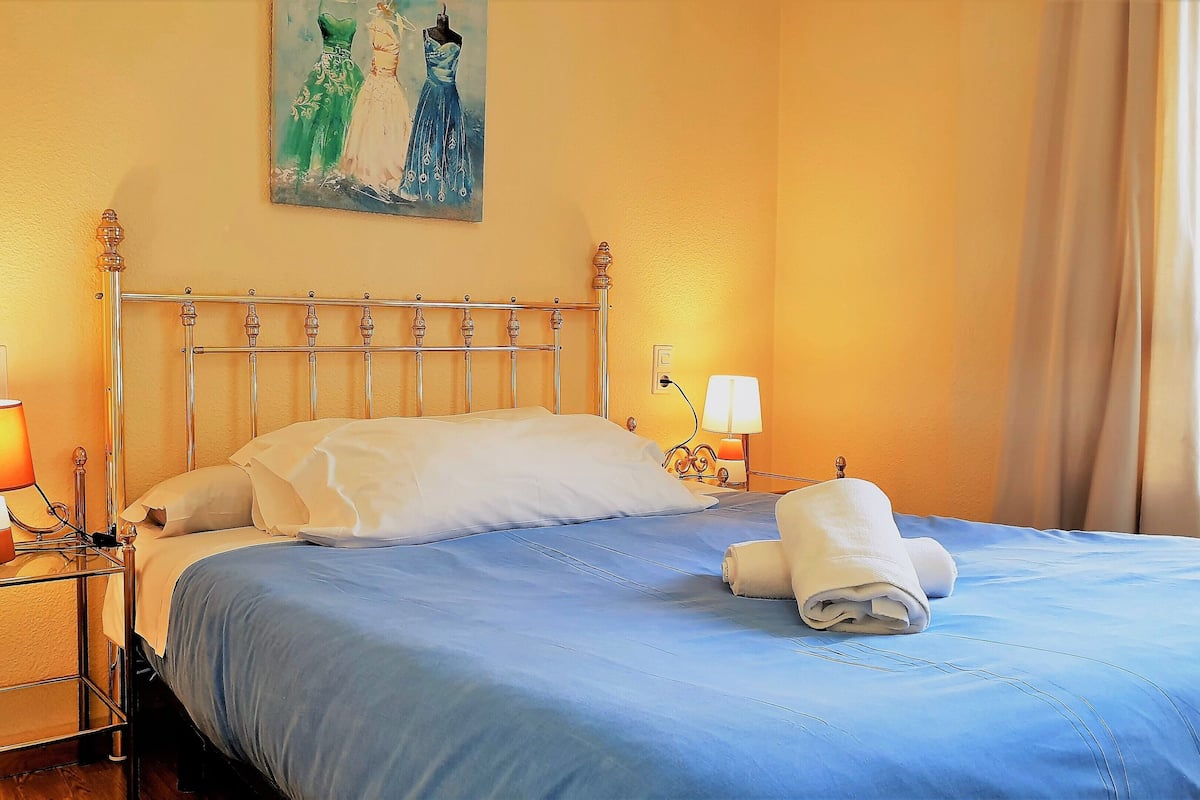 Quarto de hotel com estrutura de cama de metal, colcha azul, duas luminárias de cabeceira e um quadro na parede.