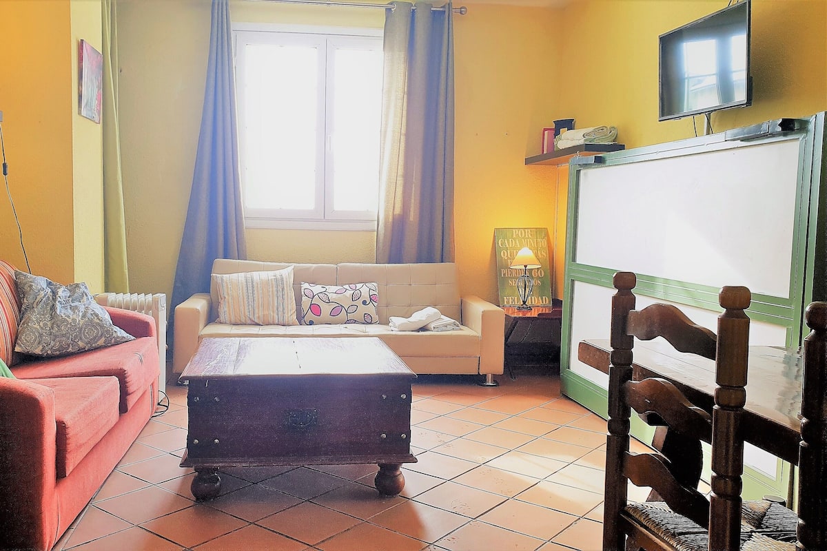 Uma sala de estar com um sofá, uma poltrona vermelha, uma mesa de centro de madeira, uma televisão e uma janela com cortinas.