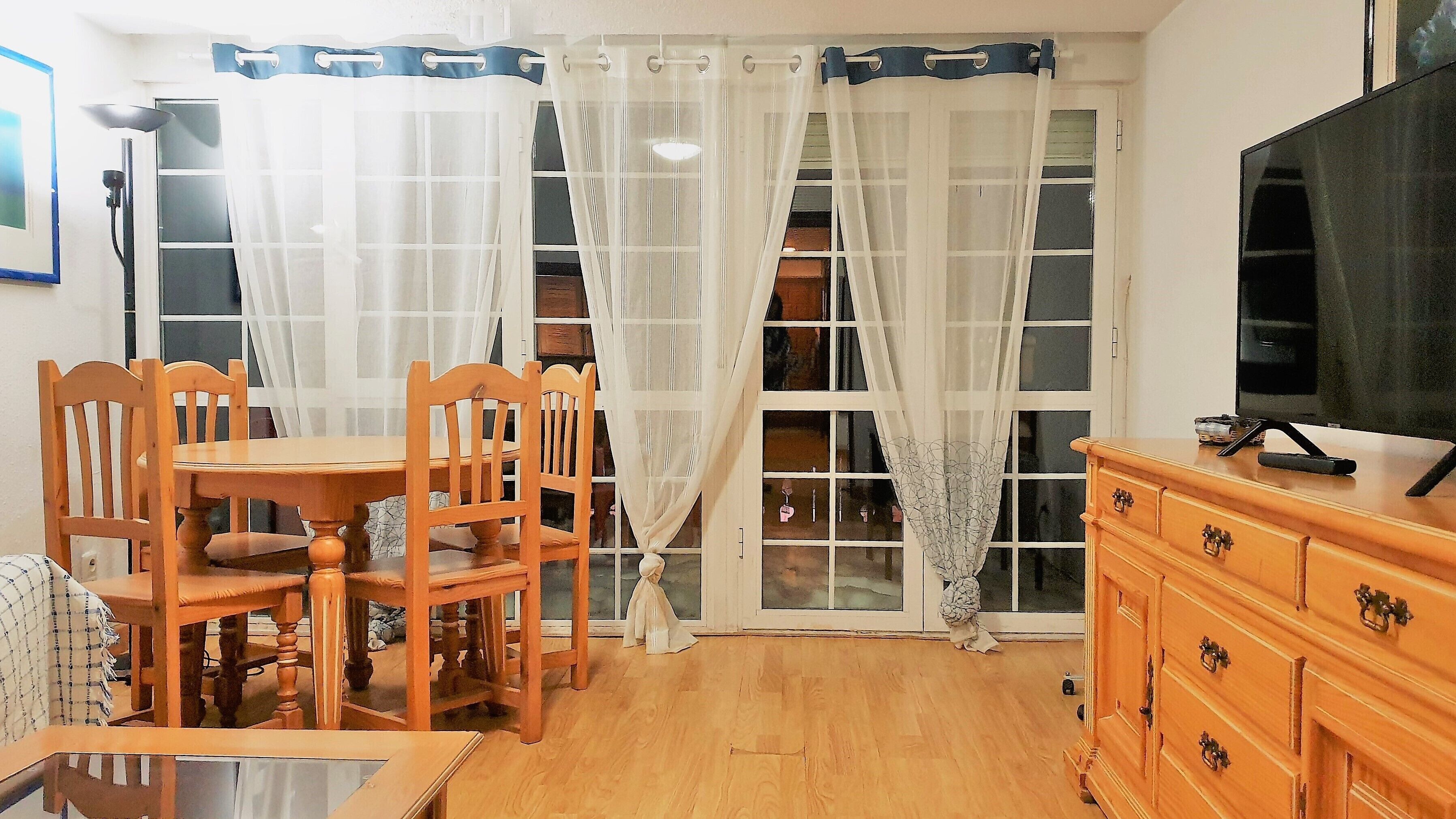 Una habitación con una mesa y sillas de madera, un televisor sobre un mueble de madera, y un ventanal con cortinas traslúcidas.