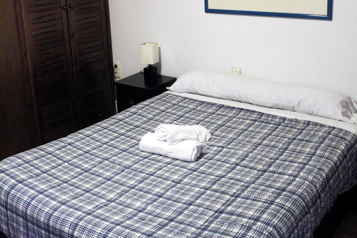 Una habitación de hotel con una cama grande, un armario, una mesita de noche con lámpara y un cuadro con una vista urbana enmarcada en la pared.