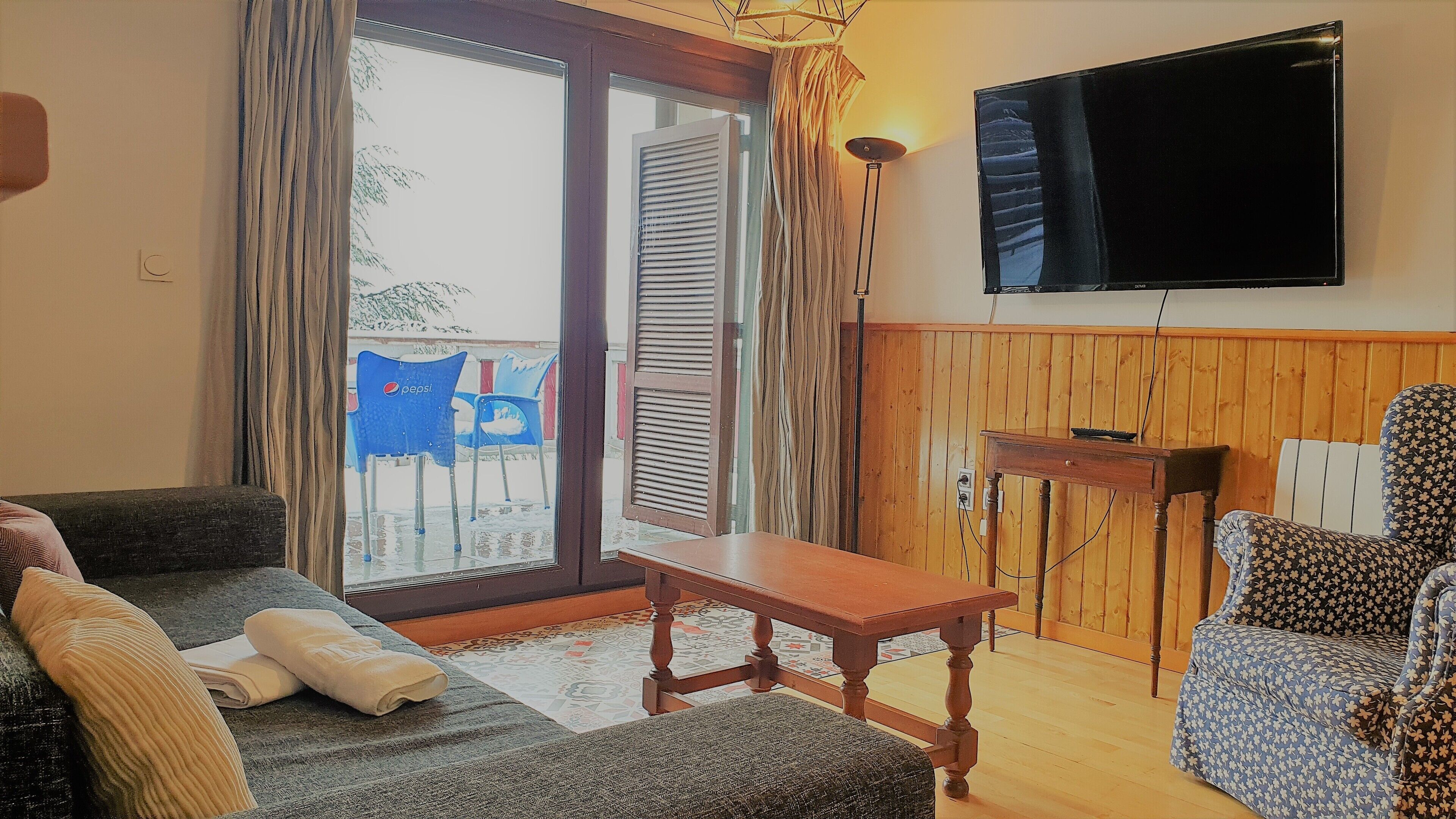Una sala de estar con un sofá, un sillón estampado, una mesa de madera y un televisor montado en la pared.
