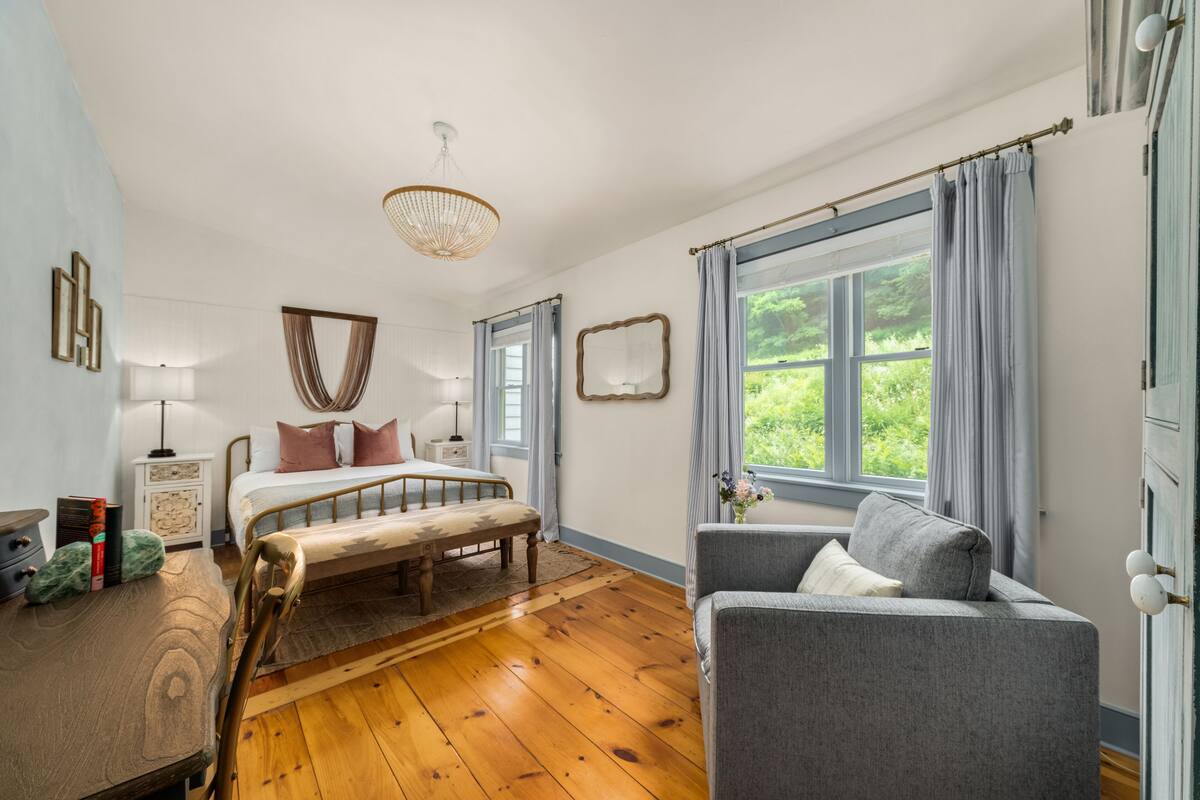 A bedroom with a wooden floor, a bed with a bench, a grey armchair, a bedside table with books, and a window with curtains.