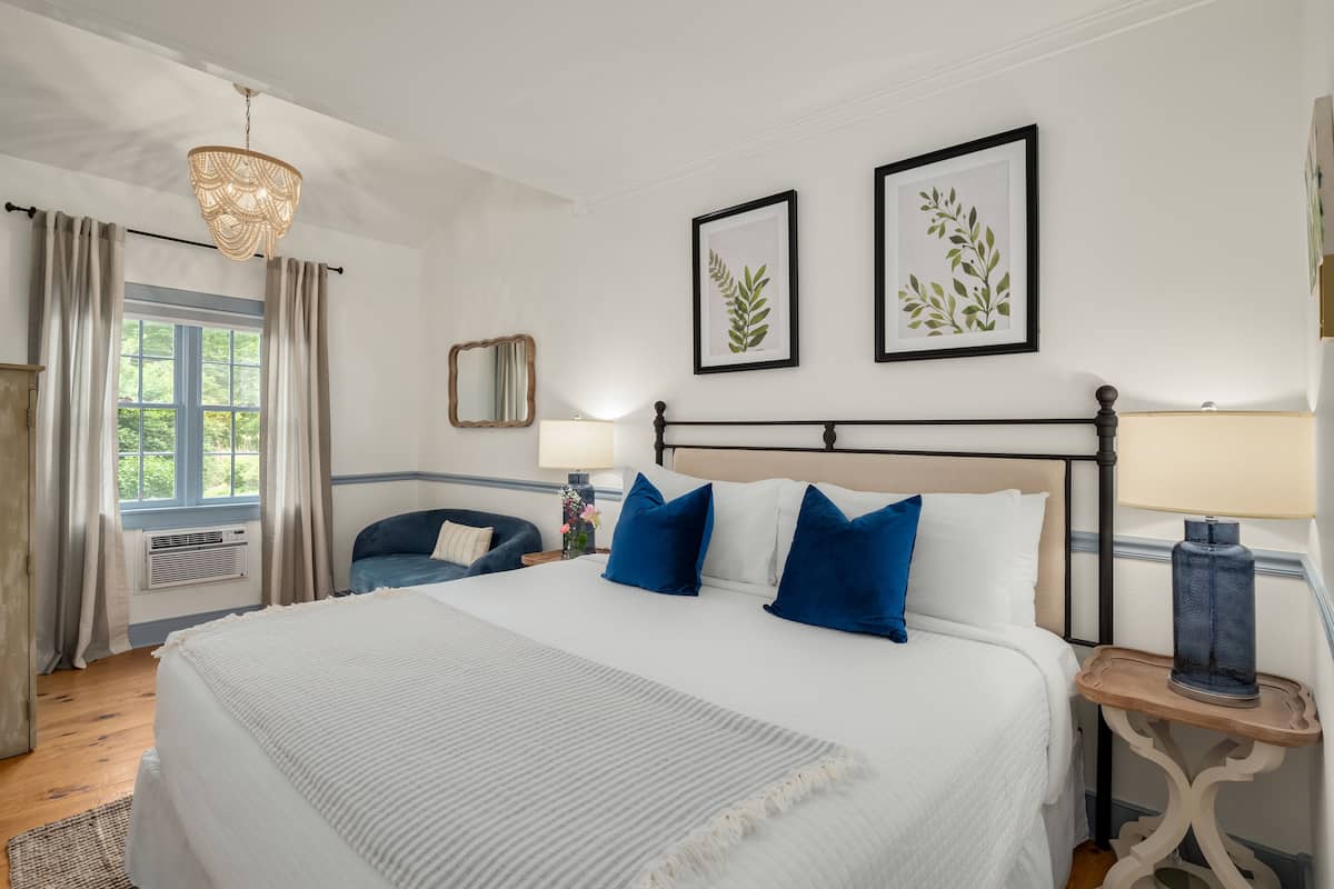 A neatly arranged bedroom with a large bed, a bedside table with a lamp, a chair, and framed botanical prints on the wall.