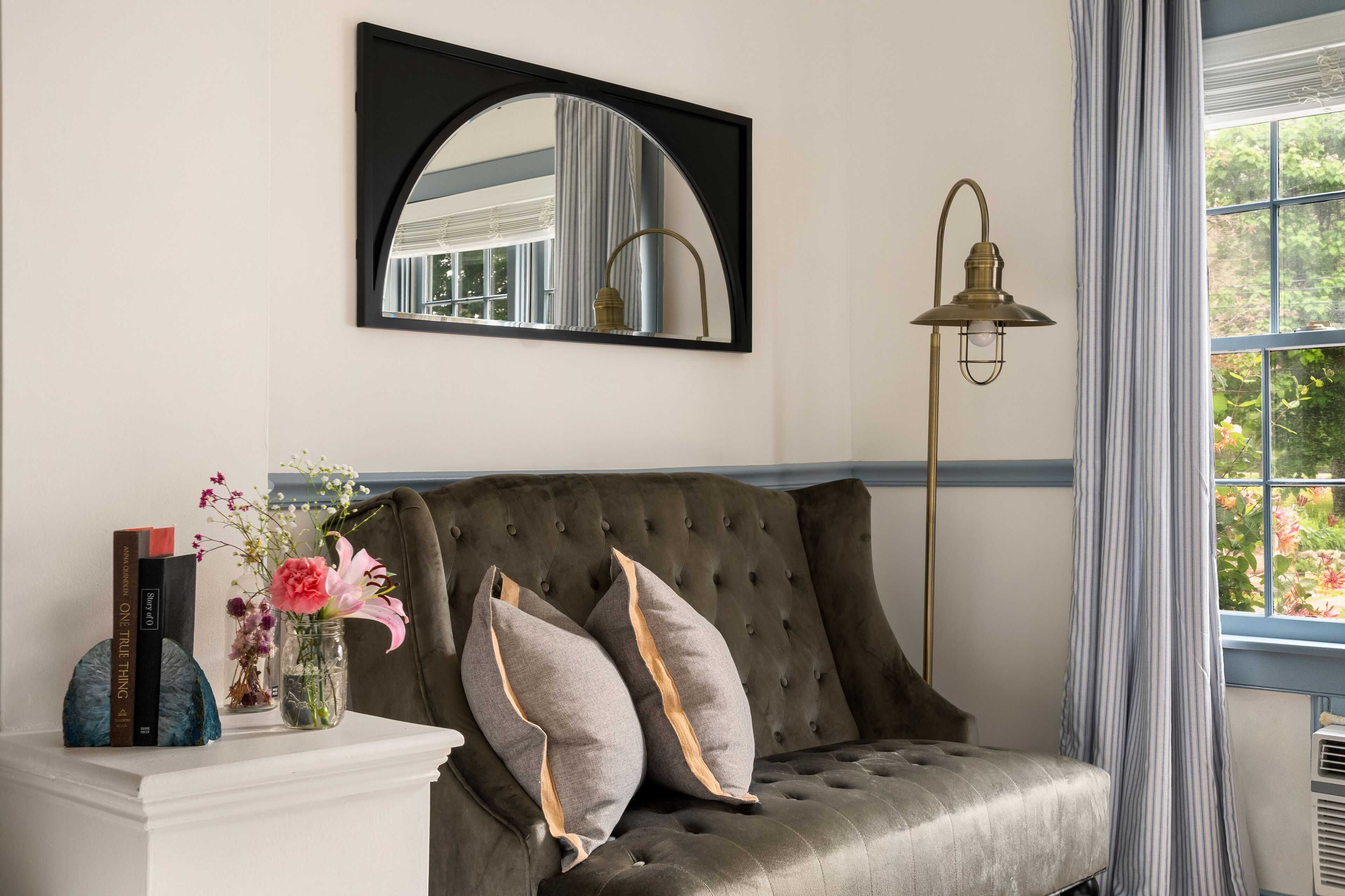 Ein Zimmer mit einer рарierten Couch, einem weißen Beistelltisch mit Büchern und einer Blumenvase, einem Spiegel, einer Stehlampe und einem Fenster mit Vorhängen.