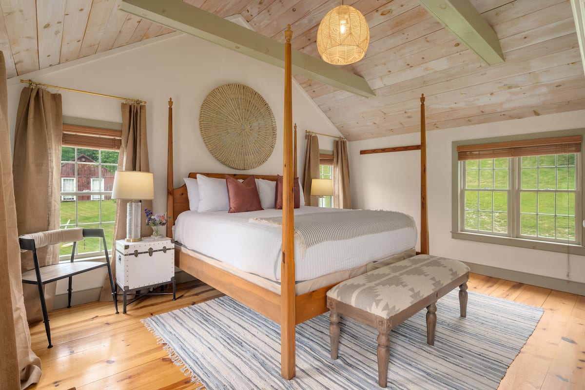A four-poster bed with a canopy, a bedside table with a lamp, a bench, and a large woven wall hanging.