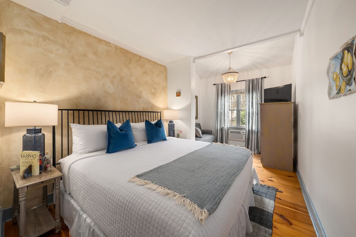 A neatly arranged bedroom with a large bed, bedside lamps, a nightstand, a grey throw blanket, and a wooden floor.