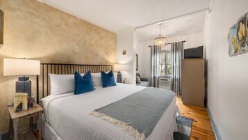 A neatly arranged bedroom with a large bed, bedside lamps, a nightstand, a grey throw blanket, and a wooden floor.