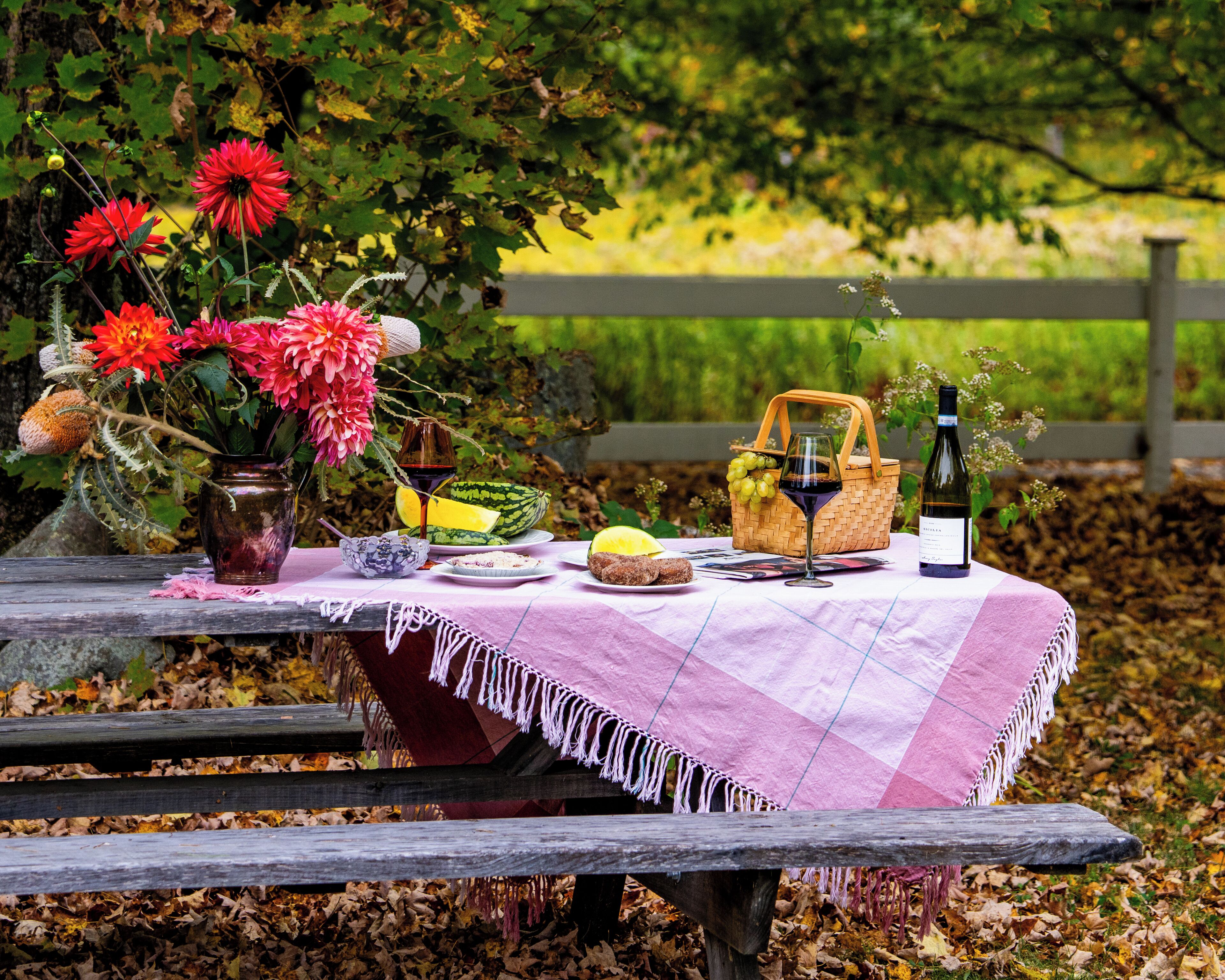Uma montagem de piquenique com uma mesa, uma cesta, uma garrafa e um vaso com flores.